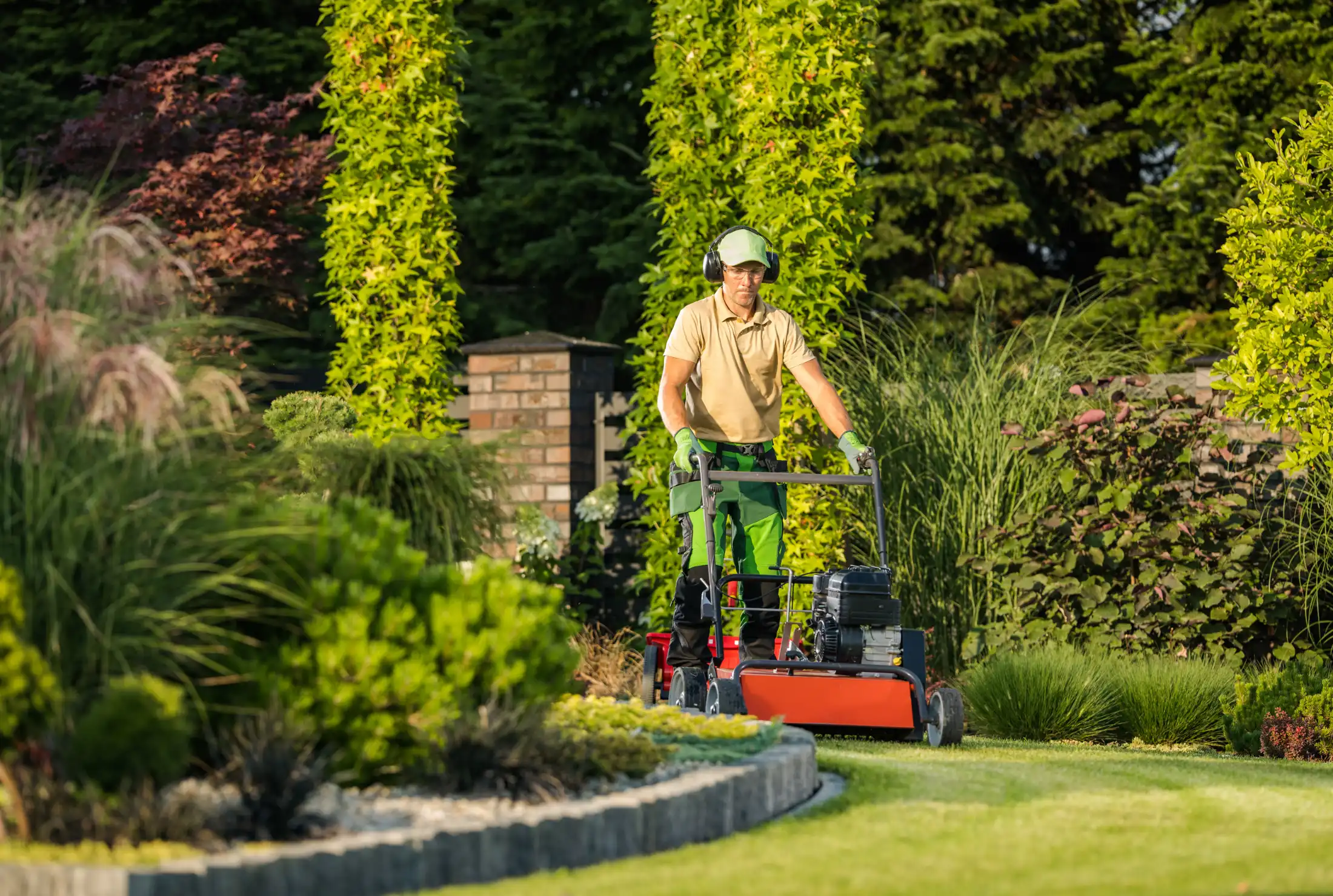 Gardener mowing a lush green lawn with a red lawnmower. Tall, leafy plants surround the area, creating a serene and tidy atmosphere.